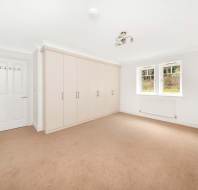 Empty room with beige carpet, white walls, and ceiling. Fitted wardrobe with six doors on the left. Two windows and radiator beneath them on the right. Door on the left wall. Ceiling light fixture.