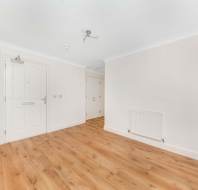 Empty room with light wood flooring, white walls, and a white front door. A radiator is mounted on the right wall, and recessed lighting fixtures are visible on the ceiling.