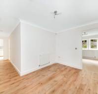 Empty room with light wood flooring, white walls, and a radiator. Open door leads to another room with windows. Ceiling lights are installed in both areas.