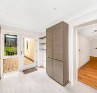 Modern kitchen with light wood cabinets, white walls, and light wooden flooring. Sliding glass door opens to a patio with a table and chairs, leading to a grassy yard.