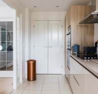 Modern kitchen with beige cabinets, a copper trash bin, double white pantry doors, built-in oven, toaster, and microwave on a light tiled floor.