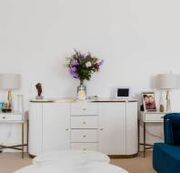 A white sideboard with flowers, two lamps, framed photos, and a blue sofa with an orange cushion in a bright living room.