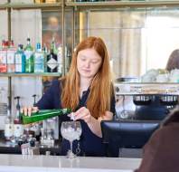 A bartender pours a green bottled drink into a glass with ice at a bar. Shelves with various bottles and a coffee machine are visible in the background.
