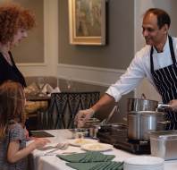 A chef in a white coat and striped apron serves food from pots on a table to a woman and a child in a dining setting. Plates and green napkins are also on the table.