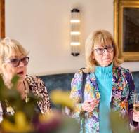 Two women with glasses and stylish outfits stand in a room. One holds a drink. They appear attentive. A painting and a light fixture are on the wall behind them.