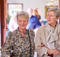 Two older women walking arm-in-arm through a hallway, with several people visible in the blurred background.