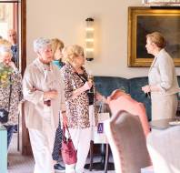 A group of older adults enter a room, one holding a drink and a shopping bag, while a woman in a blazer addresses them. The room features a framed painting and upholstered chairs.