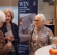 Three older women conversing near a table, with a promotional sign about winning a stay in the background.