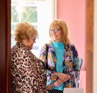 Two women with glasses and floral-patterned clothing stand inside a room, engaged in conversation near a doorway.