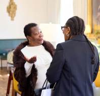 Two women having a conversation in a well-furnished room, with framed art and lamps in the background.