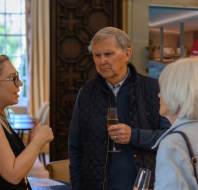 Three people stand indoors at an event. Two hold champagne glasses while engaged in conversation. A brochure table and promotional poster are visible in the background.