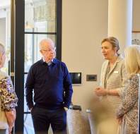 A group of four older adults, three women and one man, stand indoors having a conversation. The setting appears to be modern and well-decorated, with glass doors and columns.