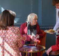 Four people sitting around a small table with drinks. One person is serving a plate of appetizers. The setting is indoors with soft light.