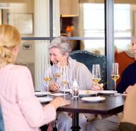 Four elderly people sit at a restaurant table, engaged in conversation, with wine glasses in front of them. The setting is bright and relaxed.