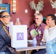 Three people sitting at a table with gift bags, engaged in conversation and holding drinks. Decor includes a blue cabinet and flowers.