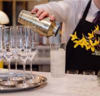 Bartender in white shirt and black apron pours liquid from a gold shaker into a tall glass with ice. Crystal wine glasses are on a tray nearby, and yellow flowers in a vase are on the counter.