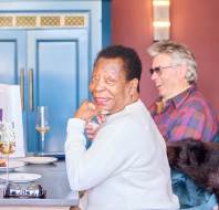 An older woman smiles at the camera while sitting at a table with a man in a plaid shirt. A blue cabinet is in the background, with flowers and drinks on the table.