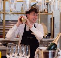 Bartender in a black apron smiles while shaking a cocktail mixer behind a bar. Hanging glasses and a champagne bottle are in the foreground.