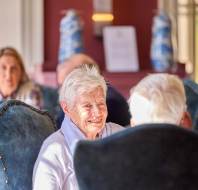 An elderly woman with short white hair smiles while seated on a blue chair in a sunlit room, with other people and decorative vases visible in the background.