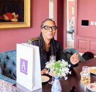 A woman seated at a table with a floral arrangement and dishes, wearing glasses and smiling. A painting hangs on a pink wall in the background.