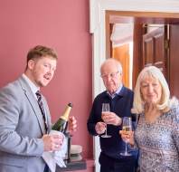 A man holding a champagne bottle, standing with an older man and woman, all holding glasses in a room with red walls and an open wooden door.