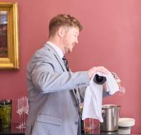 A man in a gray suit pours wine into a glass over a serving table. There are various glassware and a framed painting visible in the background.