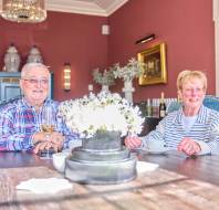 An elderly couple sits at a wooden table, smiling, with drinks in front of them. The room has pink walls, a chandelier, and decorative items. A vase with white flowers is on the table.