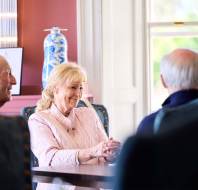 Three elderly people sit at a table in a warmly lit room. A woman in a light pink jacket smiles while talking to two men. Blue decorative vases and a window are visible in the background.