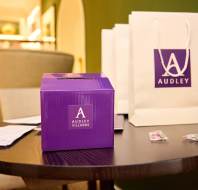 A purple box and two gift bags labeled "Audley" are on a round table. Two wrapped candies and some papers are scattered nearby. The background features a beige chair and green walls.