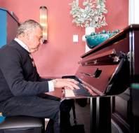 A man in a black sweater plays a piano in a room with red walls, a blue cabinet, and a decorative plant.