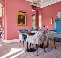 Dining room with pink walls, blue patterned chairs around a table, a blue cabinet, and a portrait on the wall. Large windows allow natural light, highlighting elegant decor and furnishings.