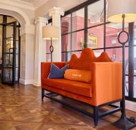 An orange velvet loveseat with pillows against a glass wall, flanked by tall lamps, in a spacious room with decorative columns and parquet flooring.