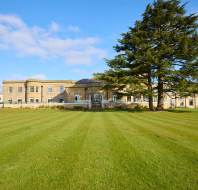 A large, historic building with a symmetrical facade stands behind a vast, well-maintained lawn and a tall tree under a clear blue sky.