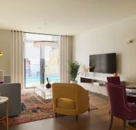 A modern living room features a gray sofa, yellow and pink chairs, a flat-screen TV, and colorful wall art. Large windows overlook a balcony with a pool view.