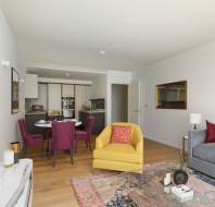 Modern living room with a yellow armchair, gray sofa, and colorful rug. A dining table with purple chairs is near a kitchen area in the background. Neutral walls feature minimalistic artwork.