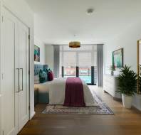 A modern bedroom with a bed, colorful throw pillows, a dresser, a large mirror, and a potted plant. A window with curtains offers a view of a building outside.