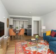 Modern living room with a blue sofa, colorful cushions, and an ornate rug. A TV is mounted on the wall, and a dining area with orange chairs is visible near the open kitchen.