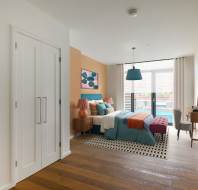 A modern bedroom with a bed, nightstands, and a desk. It features a large window, wall art, and wood flooring. Neutral and warm colors dominate the decor.