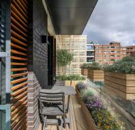 Modern apartment balcony with a wooden table, chairs, and planters filled with purple and yellow flowers. Overlooks surrounding brick buildings under a cloudy sky.
