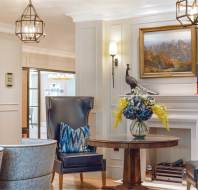 Elegant living room with two blue high-back chairs, round wooden table with floral arrangement, framed painting above a fireplace, and modern chandeliers hanging from the ceiling.