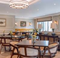 Elegant dining area with wooden tables and chairs, decorative lighting, artwork on walls, and large windows. Flower arrangements and cushions add decorative touches.