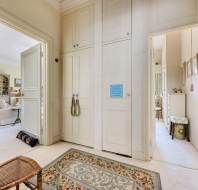 Hallway with cream-colored walls and doors, leading to a living room on the left and another room on the right. A woven rug lies on the floor, and a wooden chair is near the staircase.