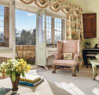 A bright, vintage-style living room with floral upholstery and a view of a garden through open French doors. A pink armchair sits next to a piano, with books and flowers on a table.