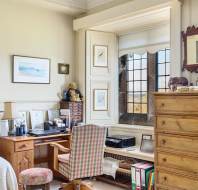 Home office with a desk by a paned window, chair, and computer. Walls are adorned with framed art and shelves hold files and collectibles. A chest of drawers and decorative mirror are nearby.