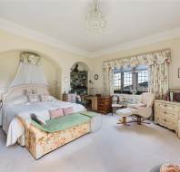 A spacious, vintage-style bedroom with floral curtains and bedding, a large bed, chandelier, armchairs, desk, and bookshelf. Large window allows plenty of natural light.