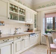 A traditional kitchen with cream cabinets, a dish rack, a small oven, and a wooden countertop. A window with a pink shade overlooks a landscape. Plates decorate the upper shelves.