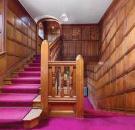 Wood-paneled staircase with red carpet and carved wood details, featuring a pendant light above and large windows on the left side.