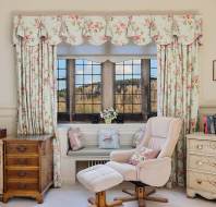 Cozy room with floral curtains framing a window view of trees. An armchair and footrest face a wooden and painted chest of drawers, next to a desk. A clock and books are visible.
