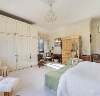 A spacious bedroom with a large bed, wooden wardrobe, cushioned chairs, and built-in white cabinets. The room is decorated with framed art and a chandelier hangs from the ceiling.