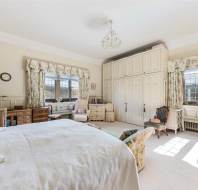 Spacious bedroom with large bed, wooden furniture, floral curtains, and built-in wardrobes. A decorative chandelier hangs from the ceiling. Sunlight streams through multiple windows.